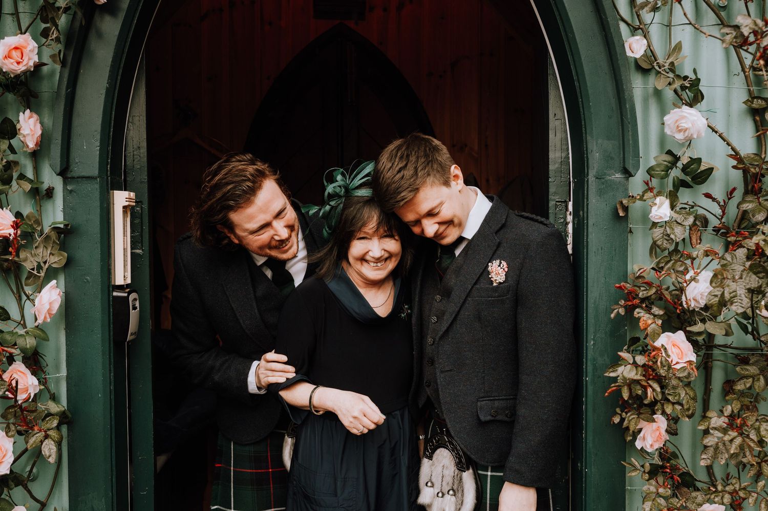 Three people laughing together in a floral-decorated green doorway.