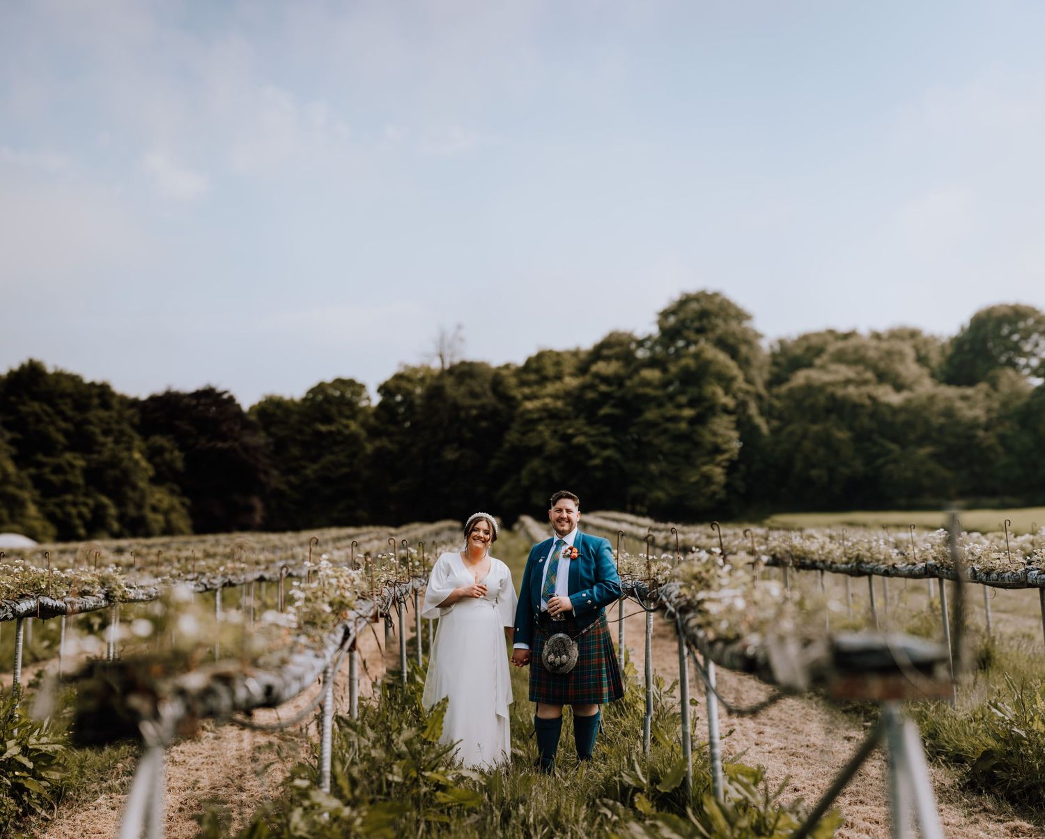Traditional Scottish wedding couple walks outdoors with rustic stone wall and greenery in background.