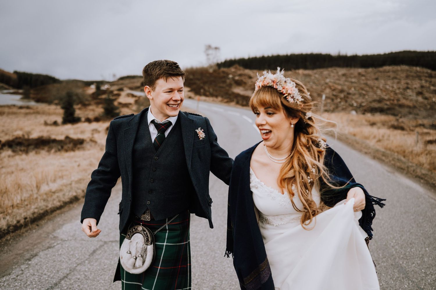 Laughing couple in Scottish wedding attire walks along rural mountain road.
