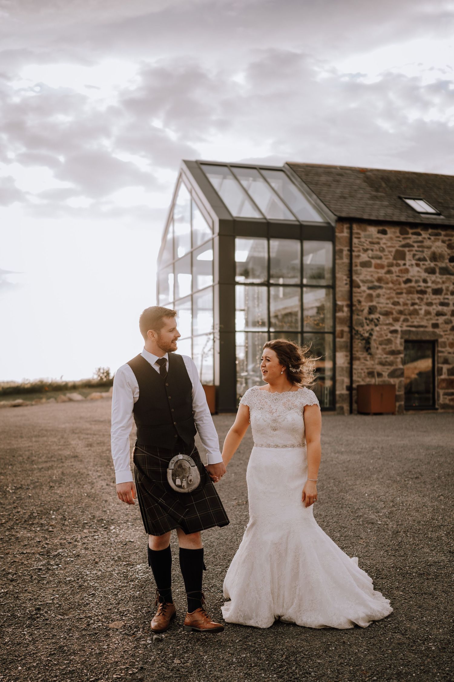 Wedding couple poses outside a rustic stone building with glass conservatory at sunset.