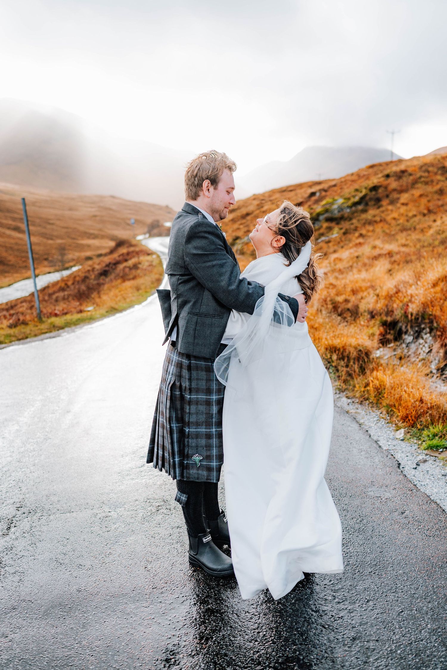 A playful moment captured on a misty Scottish road with traditional Highland dress and flowing fabric.