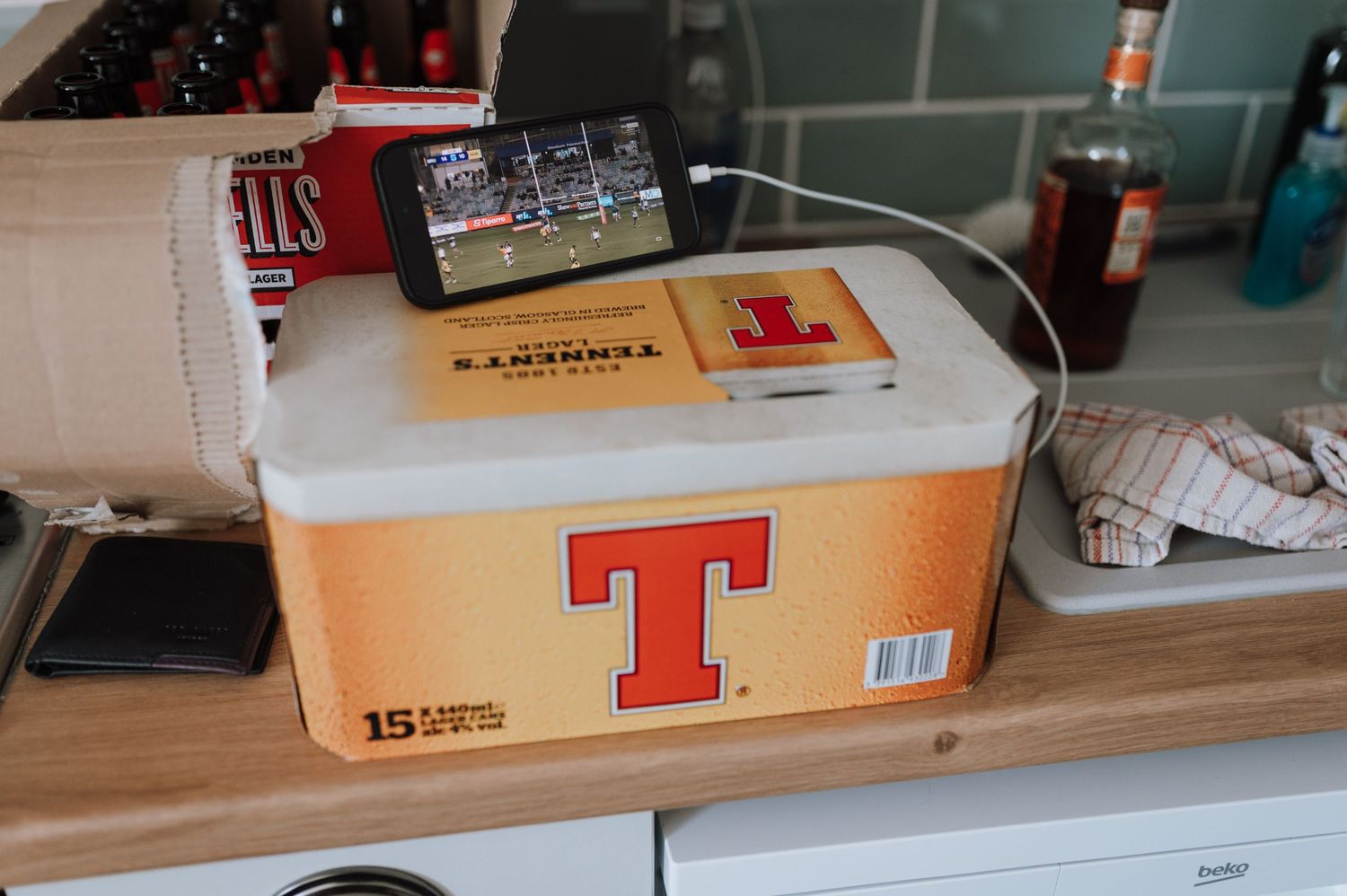 Tennent's beer case on a wooden counter with various items in background.
