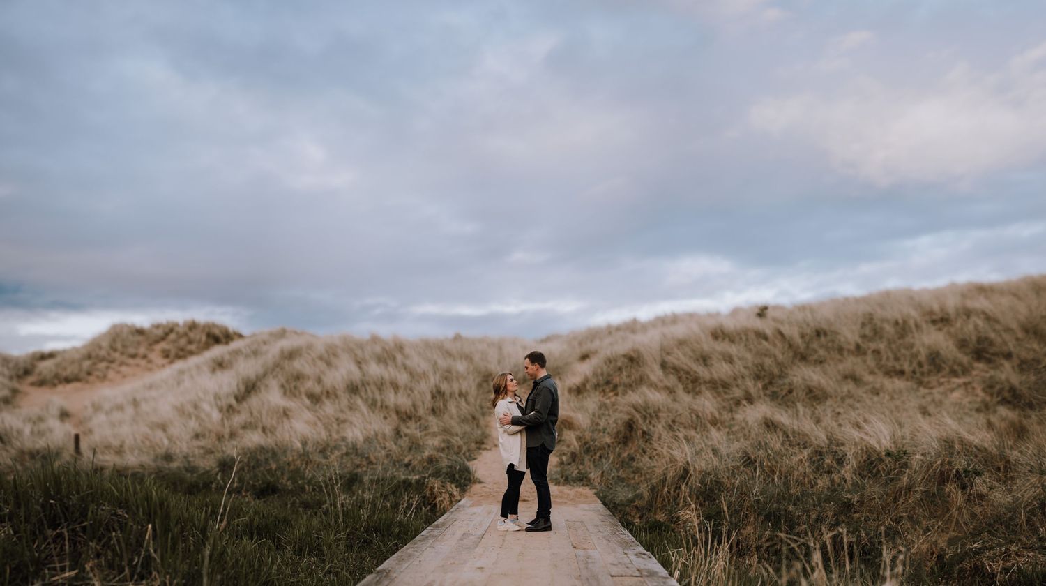 Two figures walk along a scenic pathway through sand dunes under a cloudy sky at dusk.