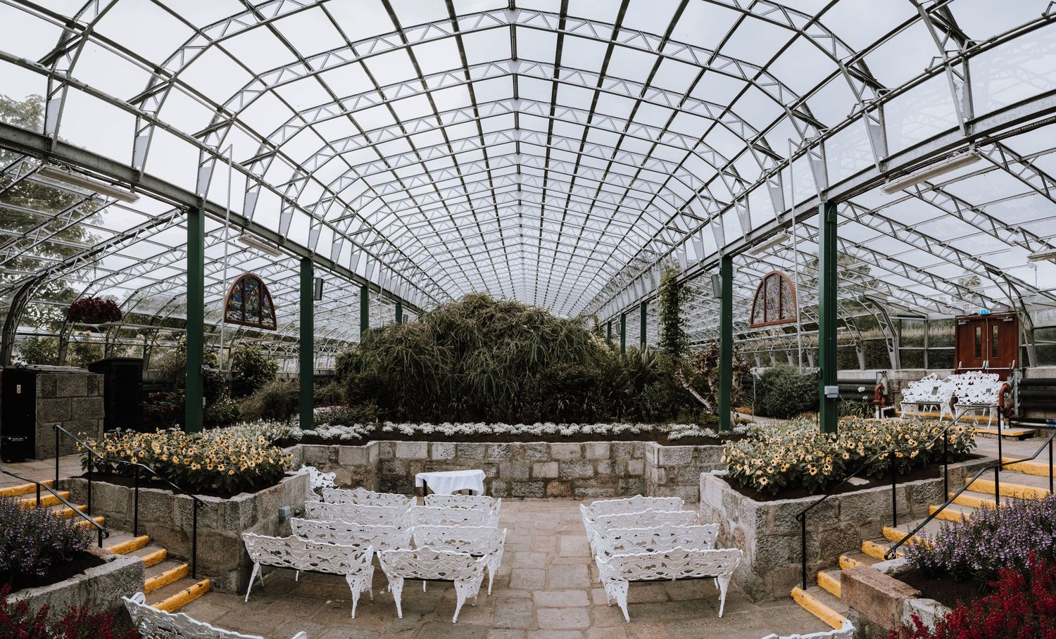 Glass-roofed conservatory set up for an elegant wedding reception with white chairs and garden surroundings.