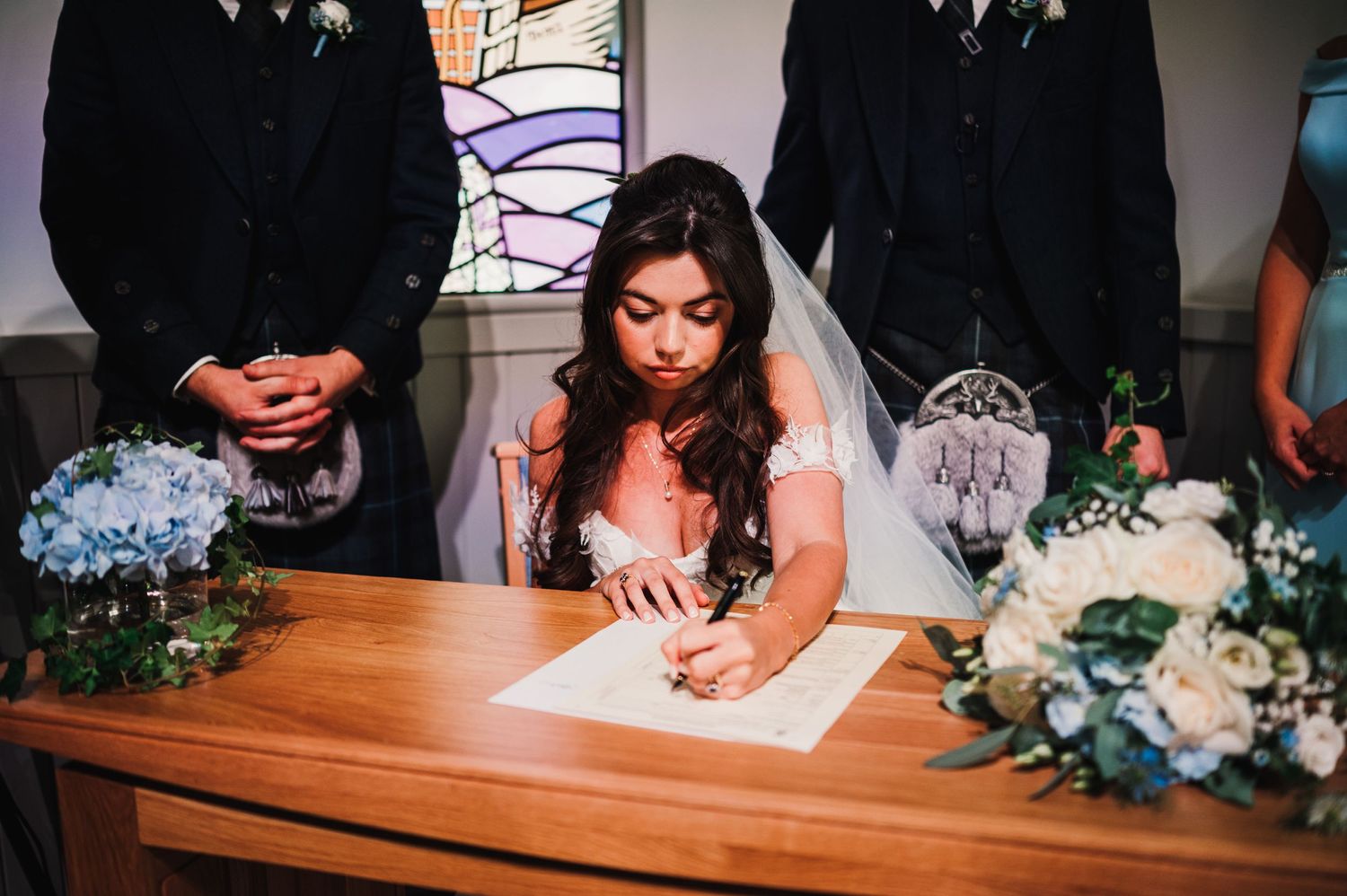 A signing ceremony at a wedding with blue hydrangea bouquets and stained glass windows.