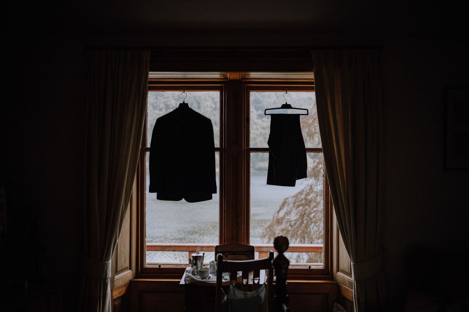 Two formal outfits hanging in silhouette against a bright window.