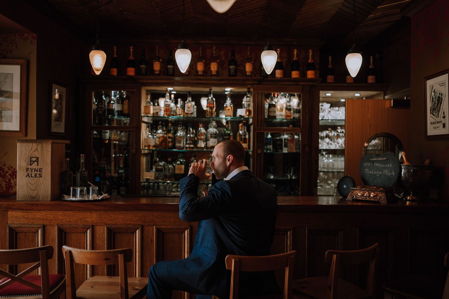 Person sitting alone at vintage bar with illuminated pendant lights and glass display.