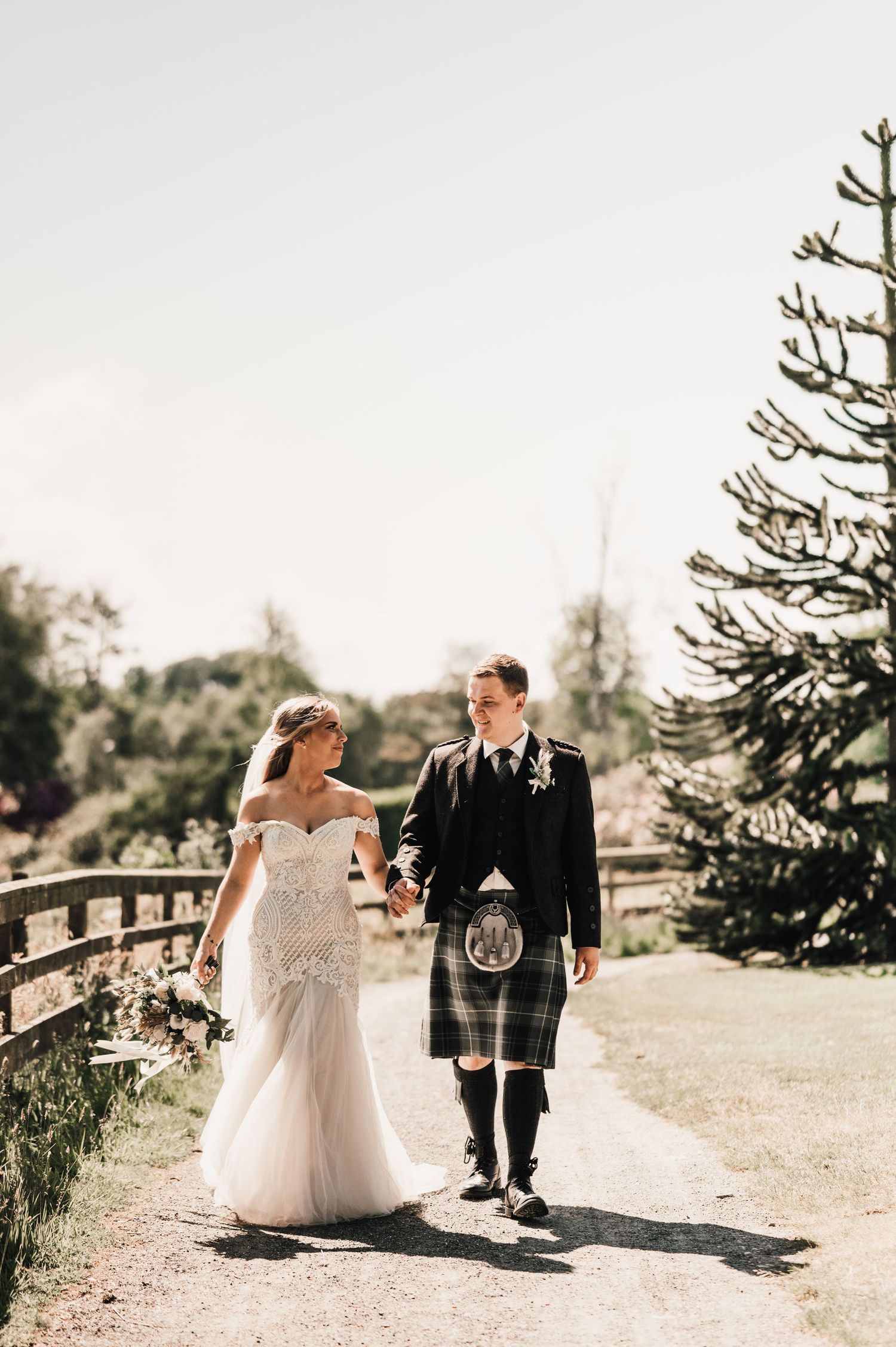 A wedding couple walks together on a garden path with the groom wearing a traditional Scottish kilt.