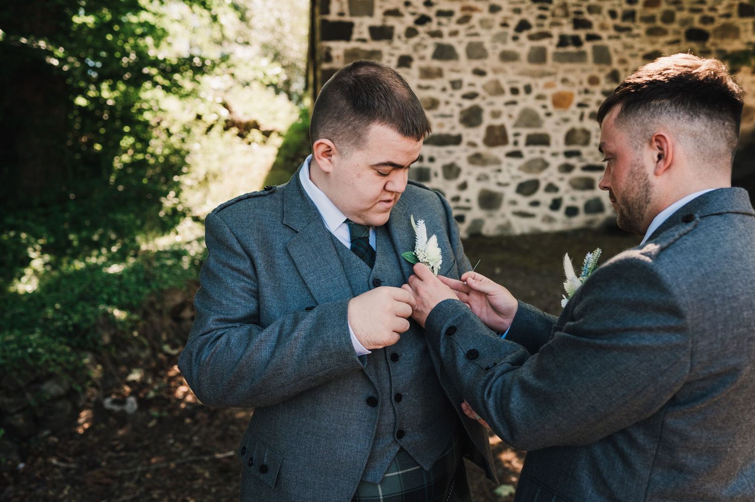 Two people in formal suits share a close moment while preparing boutonnières outdoors.