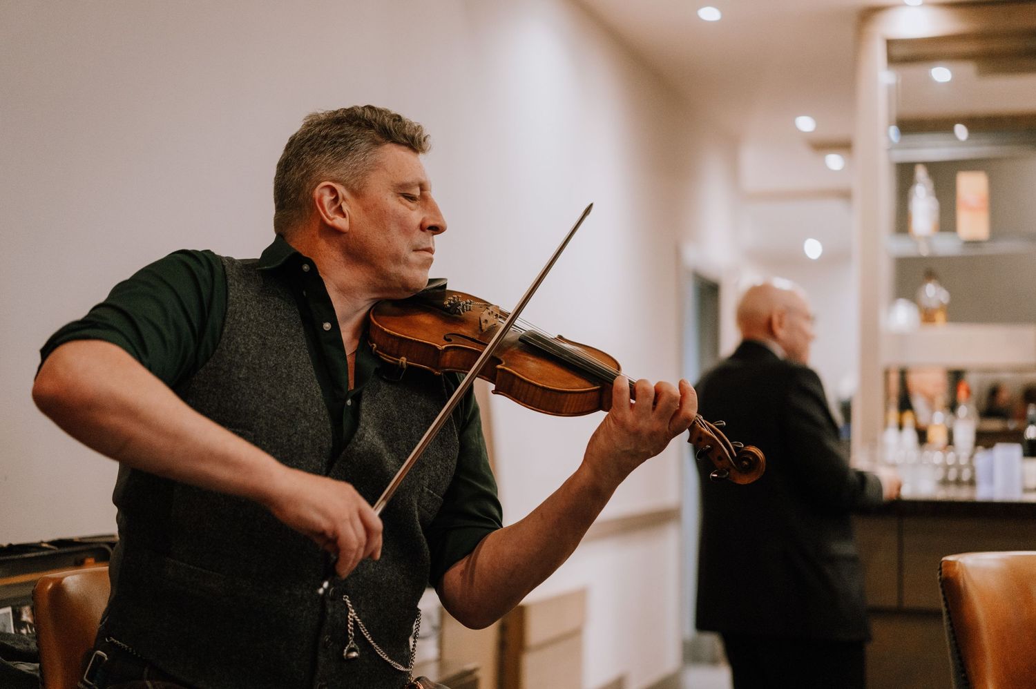A violinist performs in formal attire at an indoor venue with someone visible in the background.
