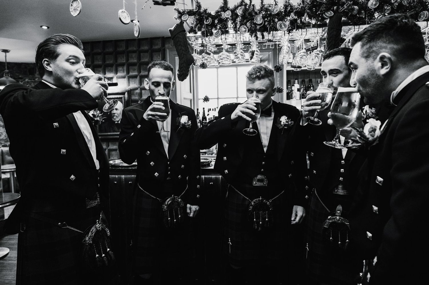 Groomsmen in black formal wear sharing drinks and conversation at a wedding celebration.