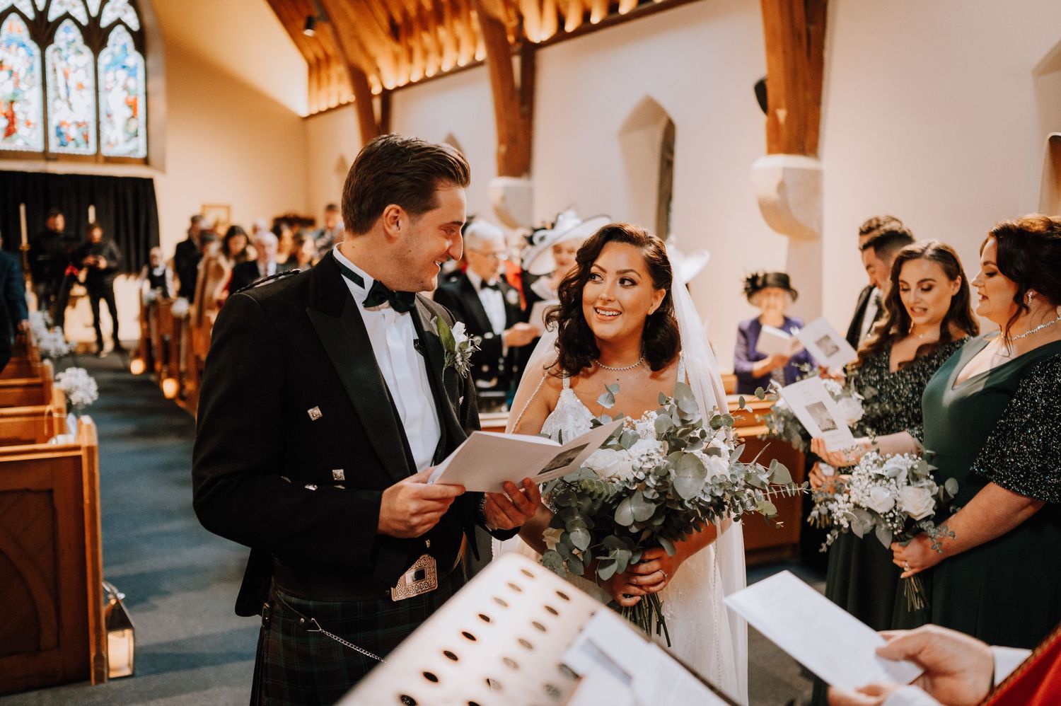A couple in traditional Scottish wedding attire reads from a program during their church ceremony.