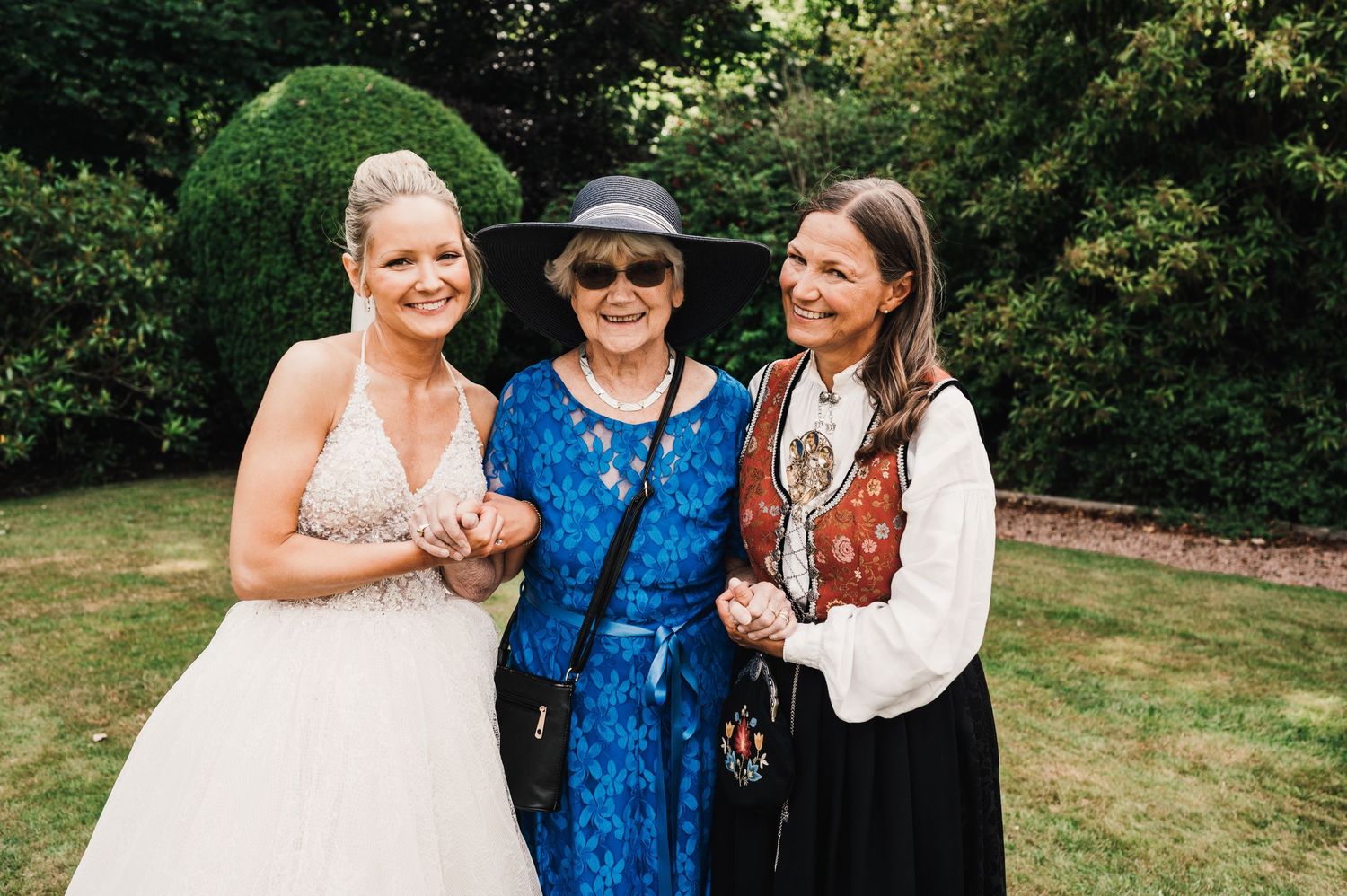 Three people in wedding attire pose outdoors near a manicured green bush on a sunny day.