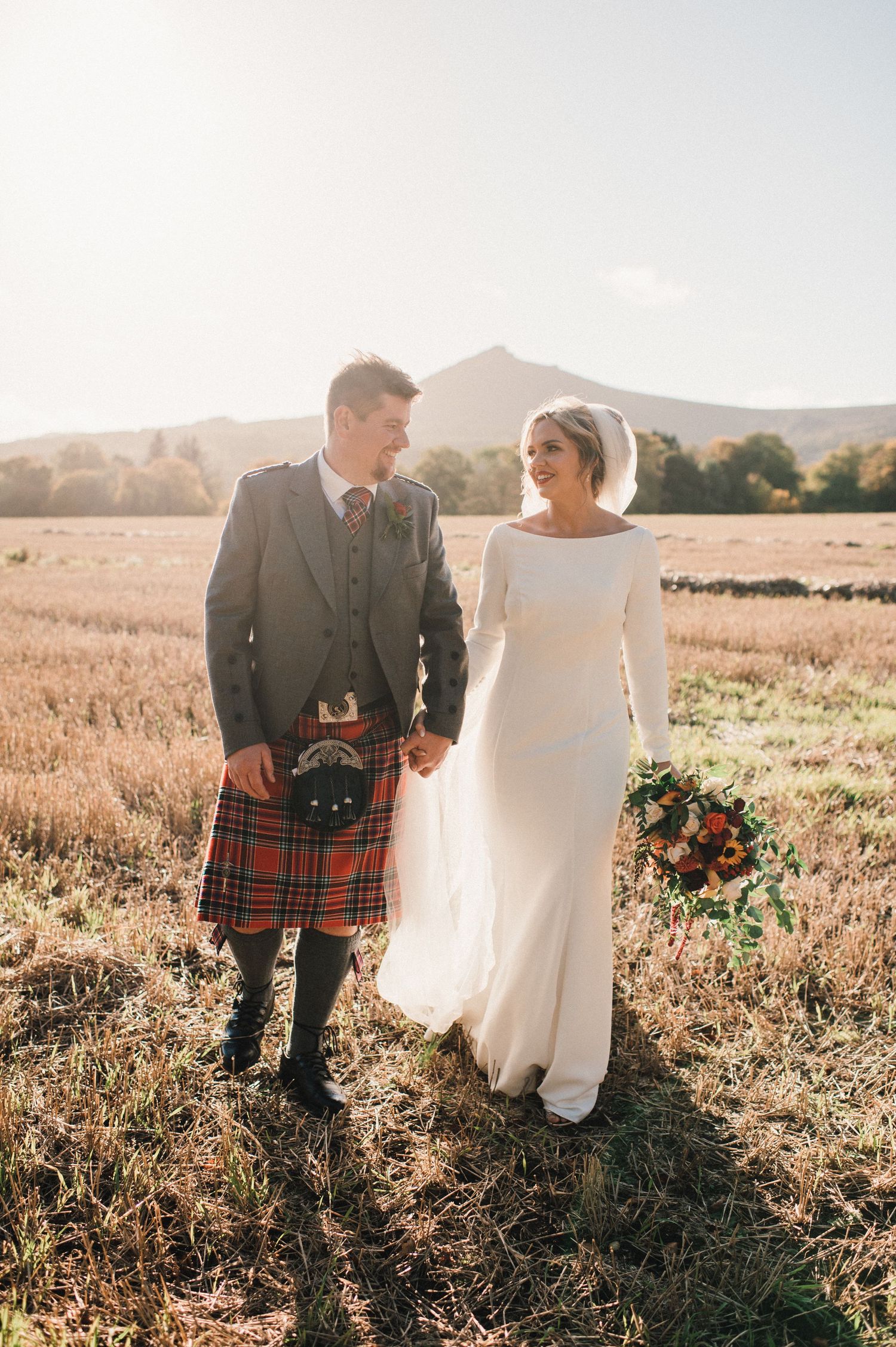Couple in Scottish wedding attire standing in golden field during sunset.
