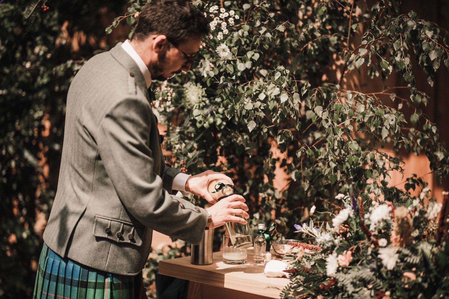 Person in traditional Scottish attire signs documents at rustic wooden table with floral decor.
