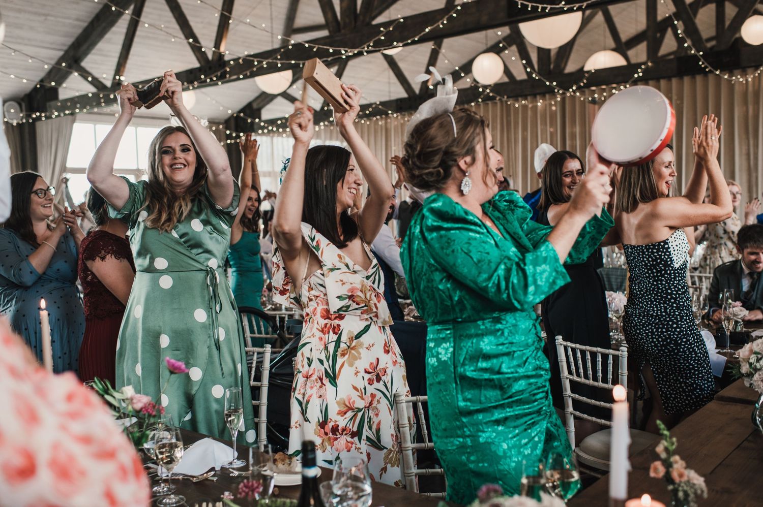 Guests in green and floral dresses dance and celebrate at a wedding reception with fairy lights.