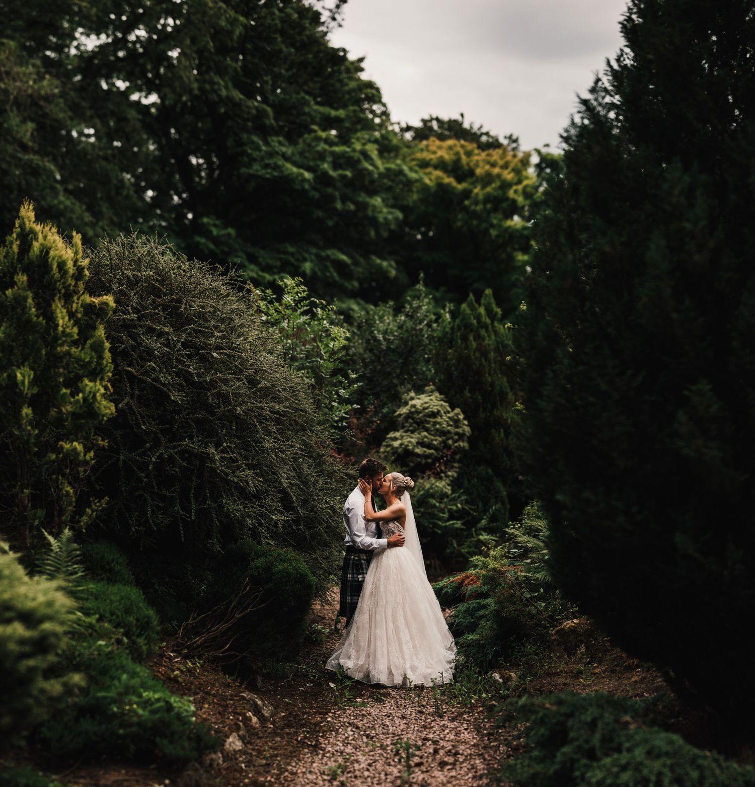A romantic wedding photo of a couple embracing on a garden path surrounded by lush greenery.