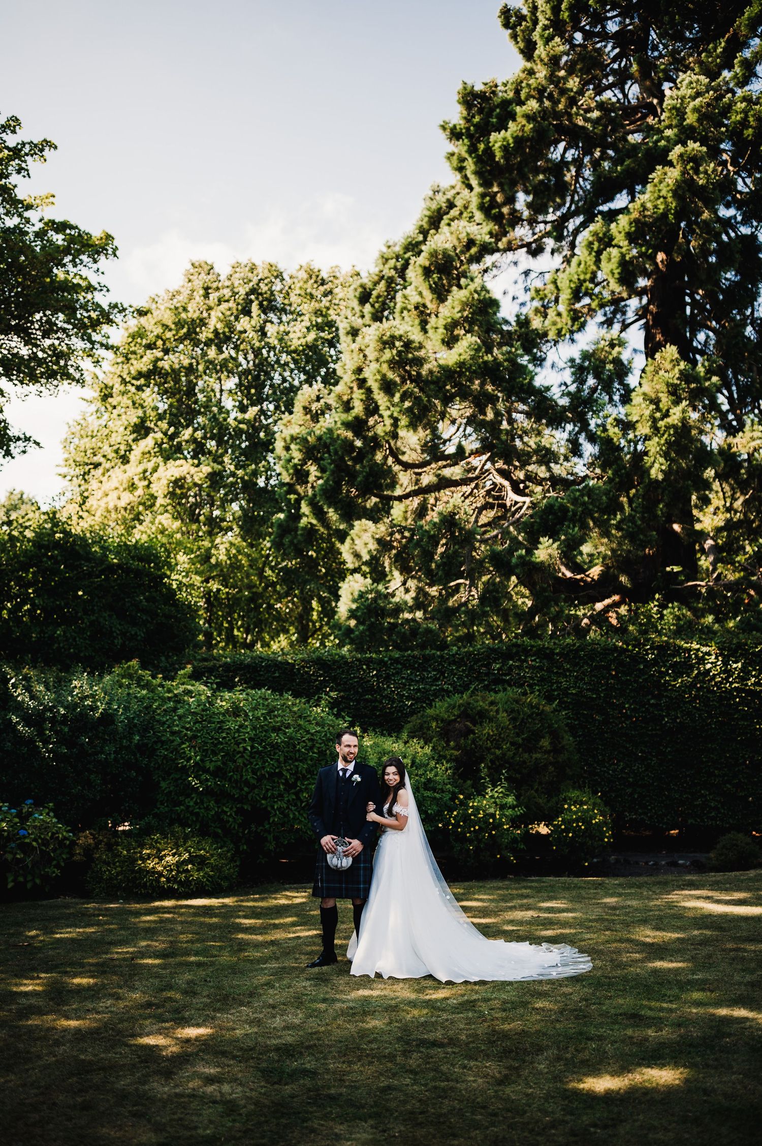 Sunlit wedding portrait in lush garden setting with towering trees creating natural backdrop.