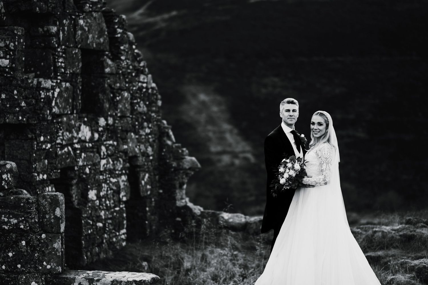 Dramatic black and white wedding portrait featuring stone ruins and moody sky.