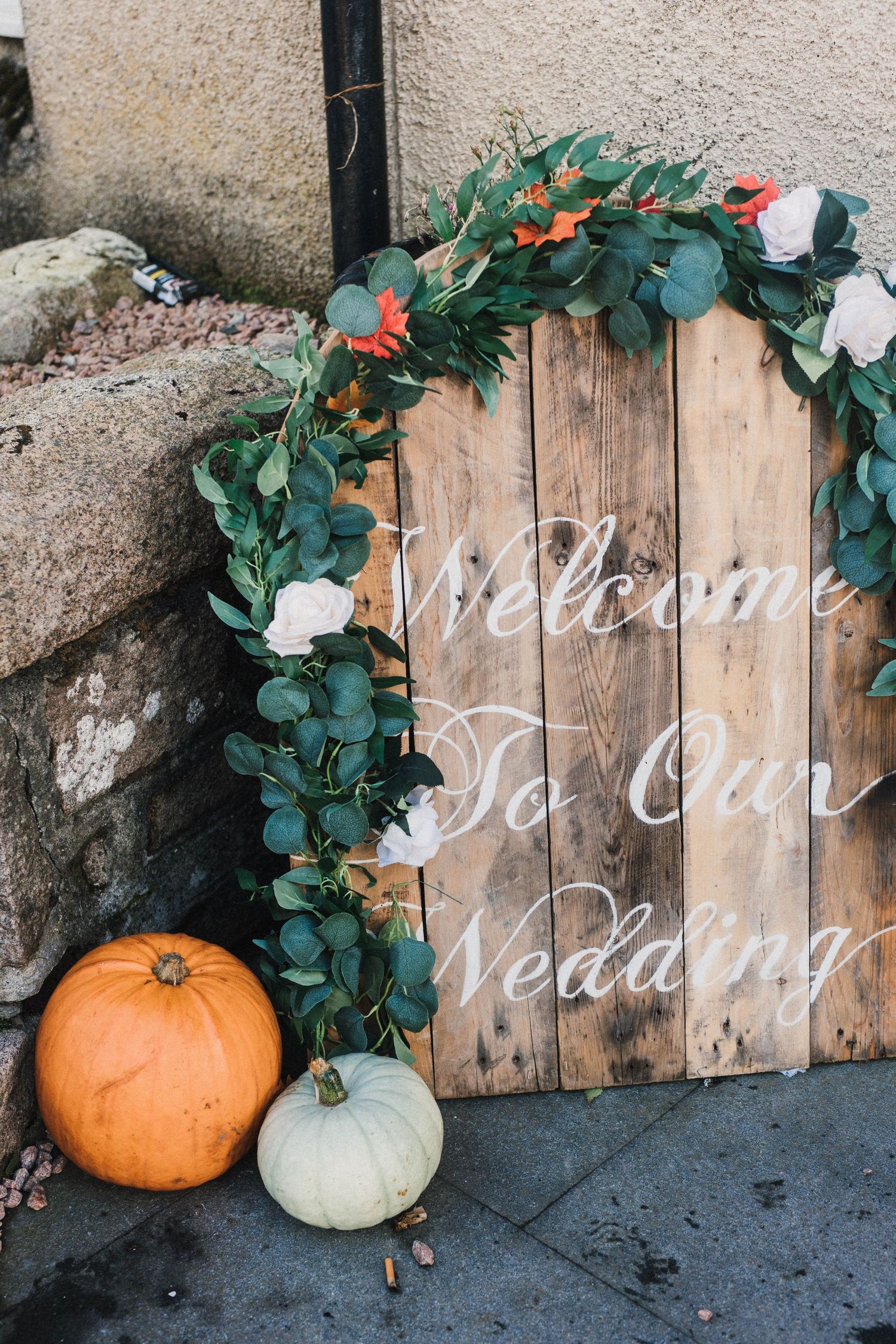 Rustic wooden wedding welcome sign decorated with eucalyptus garland and fall pumpkins.