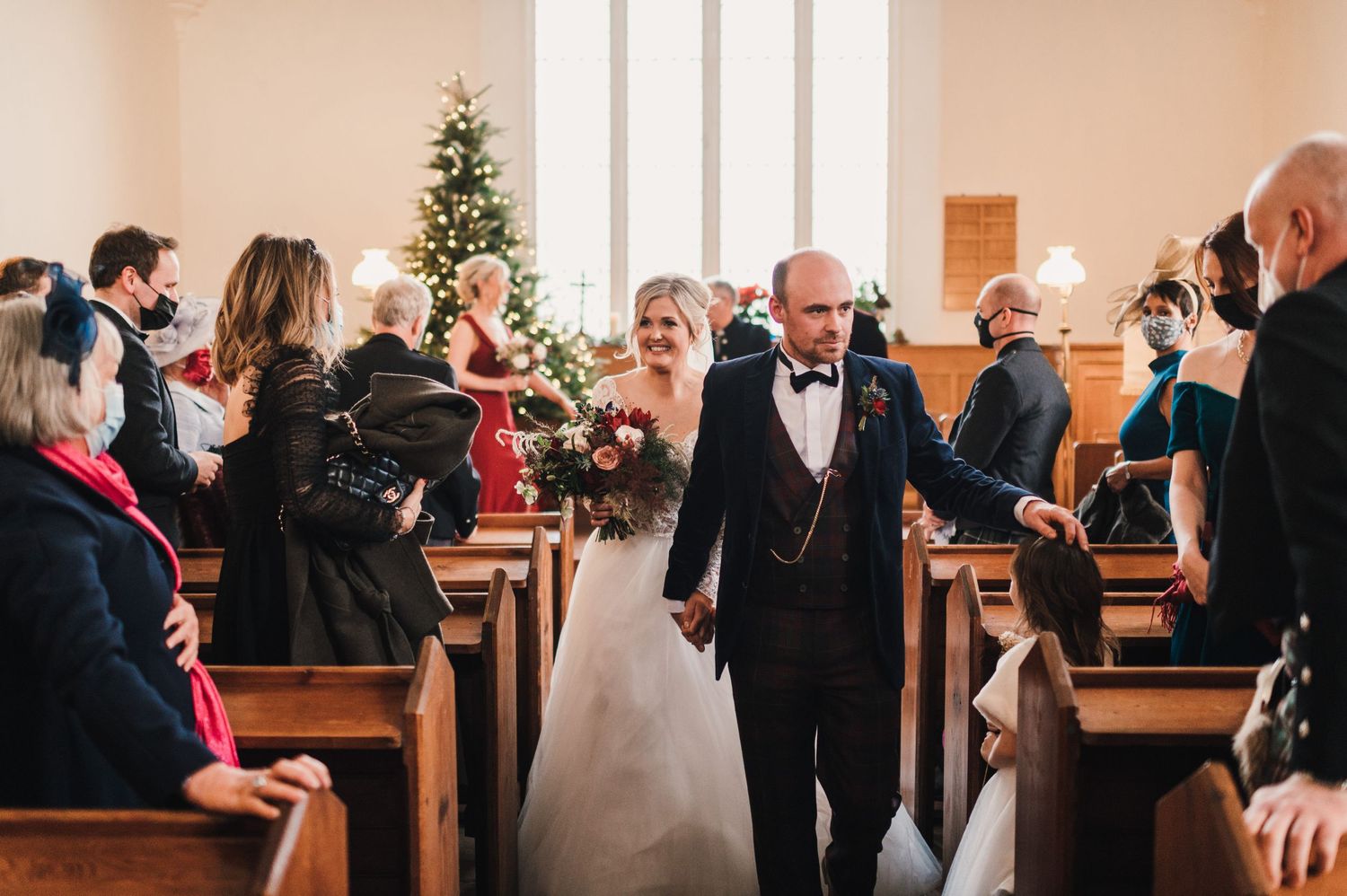 Newlyweds walking down church aisle while guests celebrate from wooden pews.