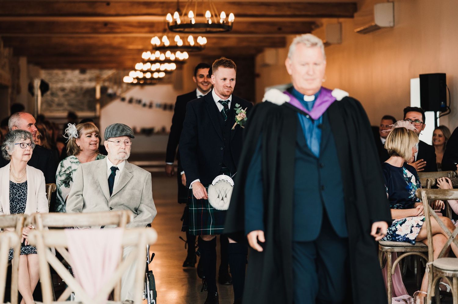 Wedding ceremony processional in an indoor venue with wooden beams and elegant chandeliers overhead.