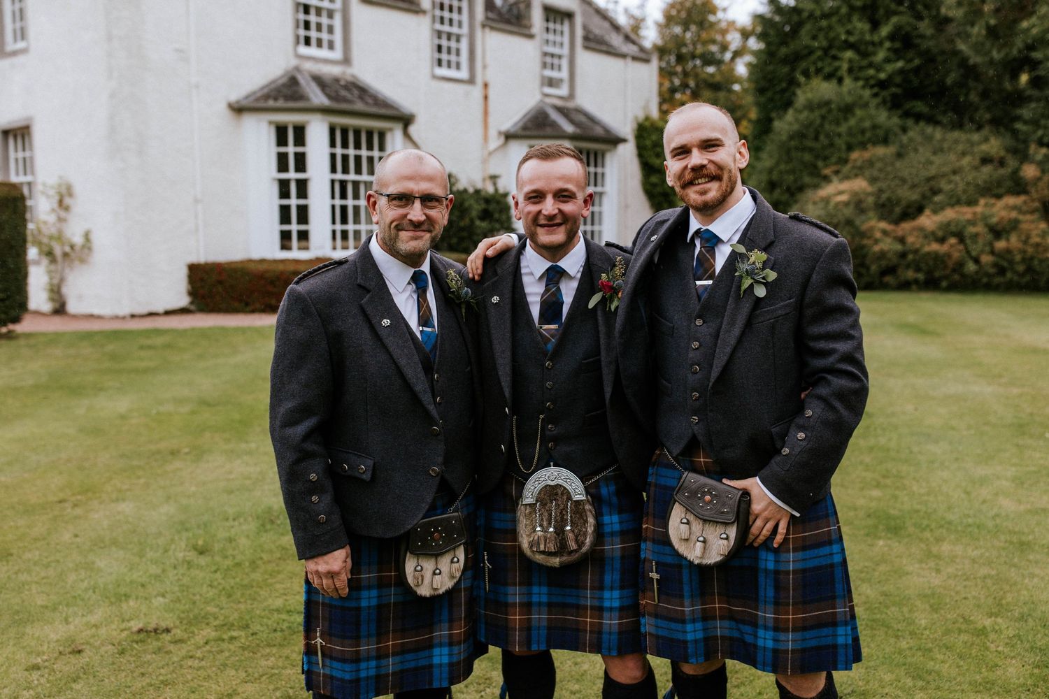 Group of men in kilts and suits stand together on manicured grounds near a white building.