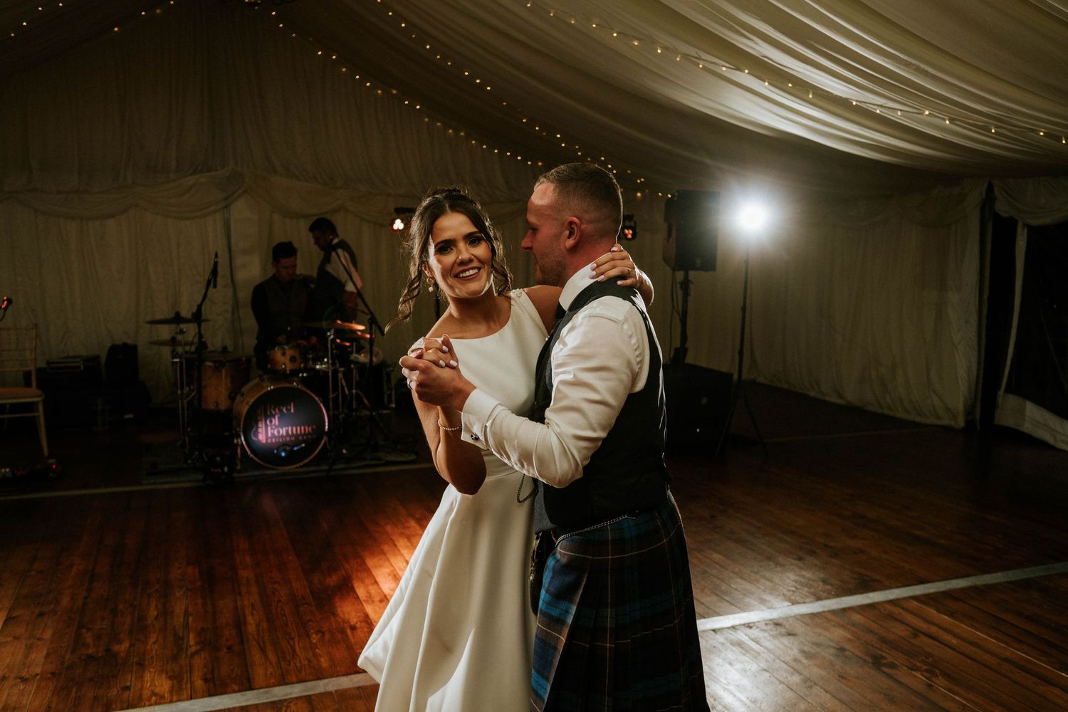 Wedding guests dancing and celebrating on a wooden dance floor under white tent draping.