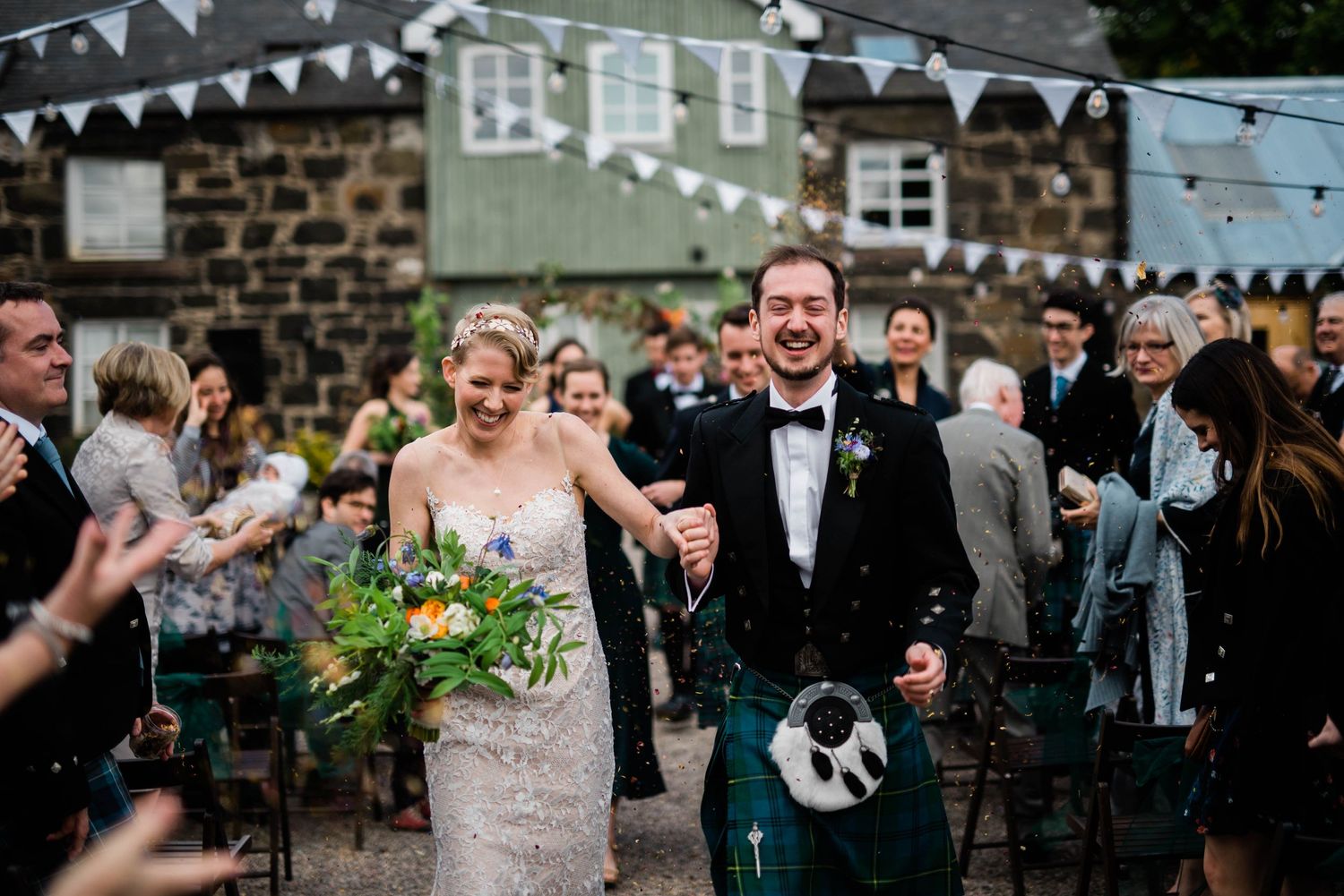 A joyful outdoor wedding celebration with Scottish kilts and white bunting at a stone building venue.