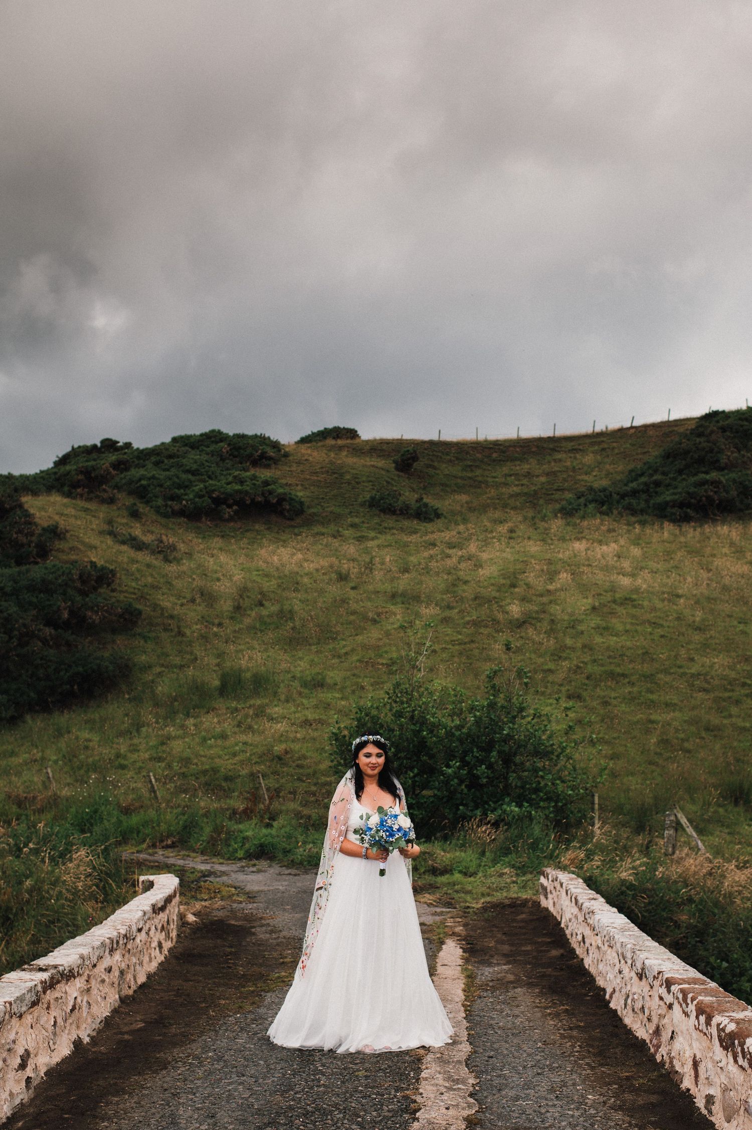A bride in white stands alone on a stone bridge against moody Scottish hills and cloudy skies.