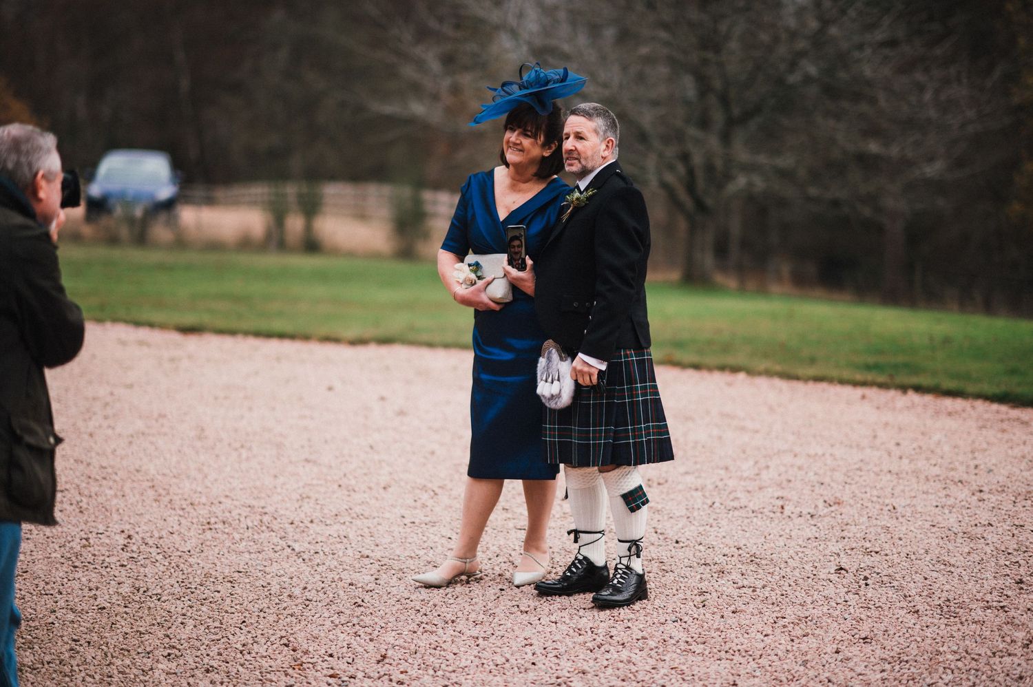 Wedding guests in traditional Scottish and formal attire converse on gravel path.