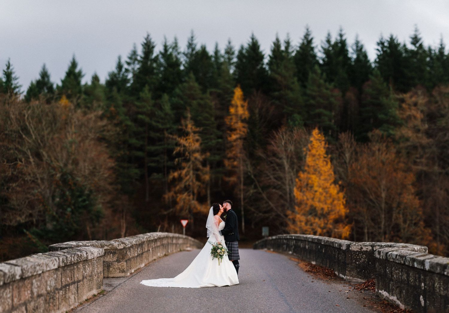Bride in flowing white gown stands on stone bridge with autumn forest backdrop.