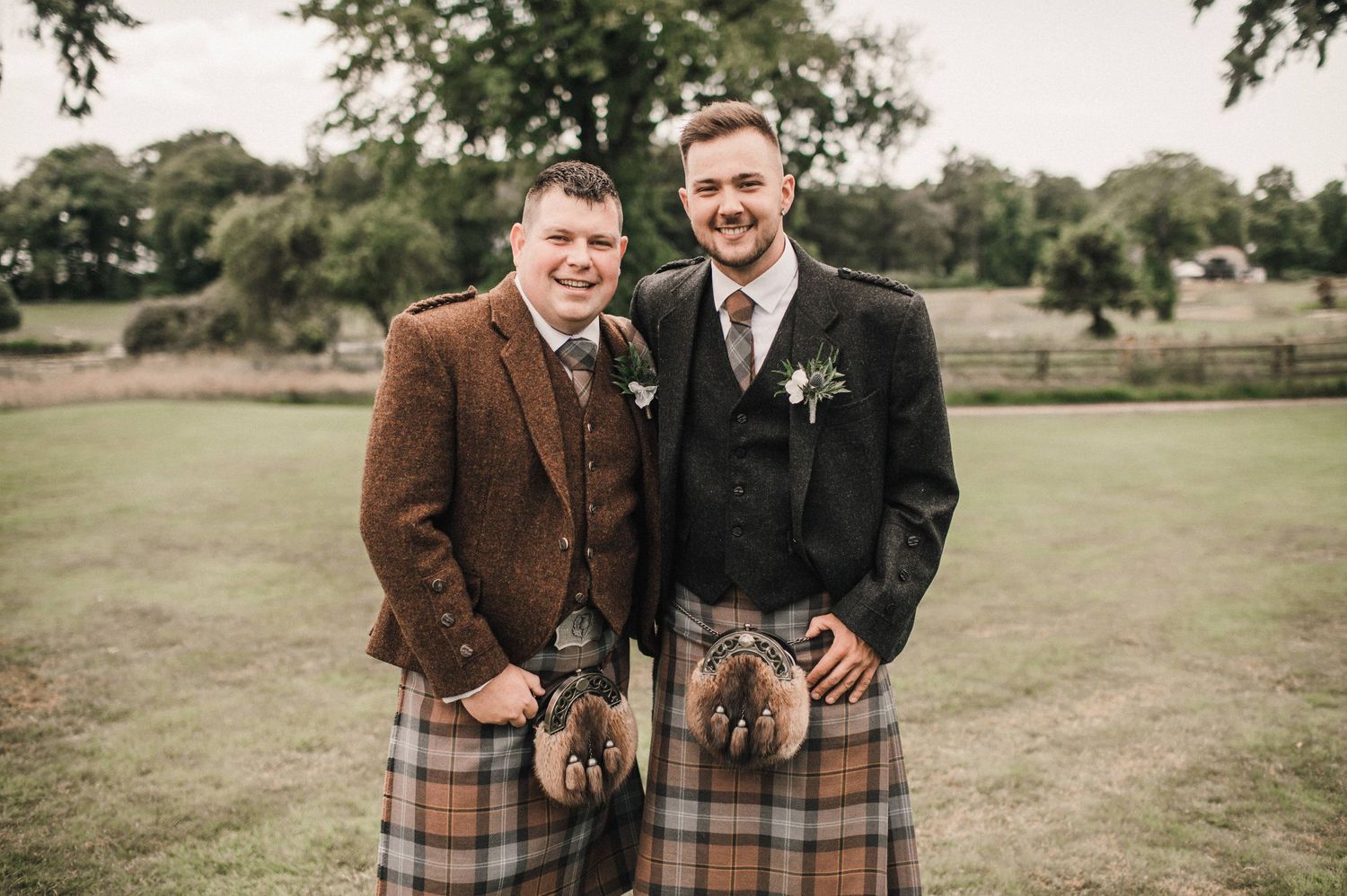 Two men in traditional Scottish wedding attire with kilts pose together outdoors.