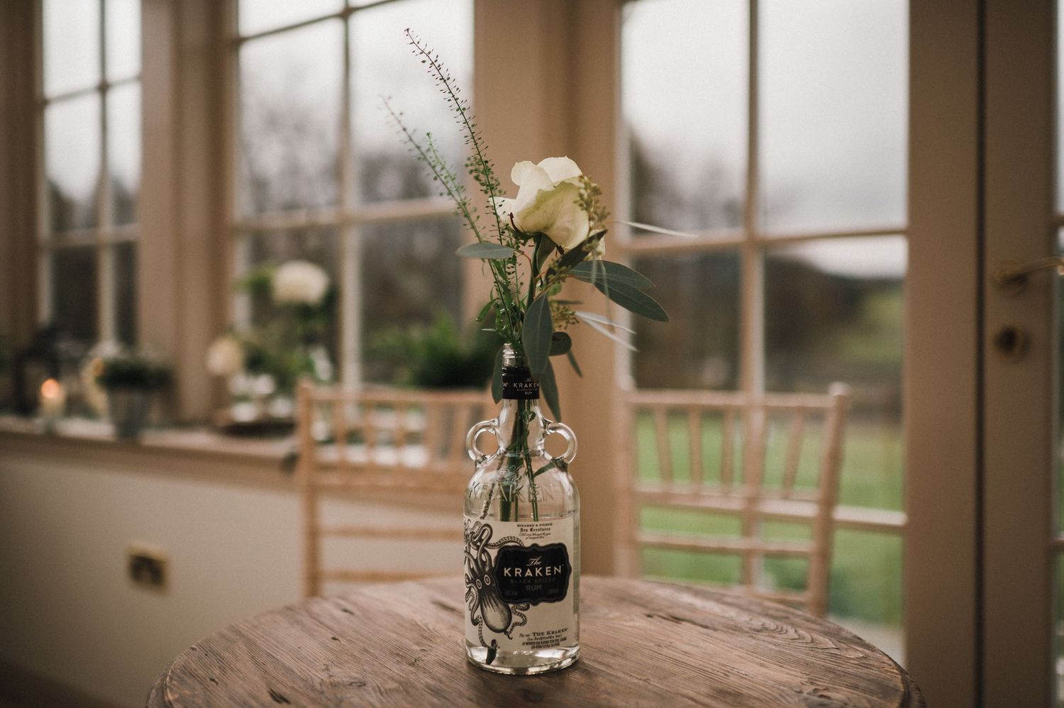 Simple floral arrangement in glass bottle vase sitting on wooden table near window.