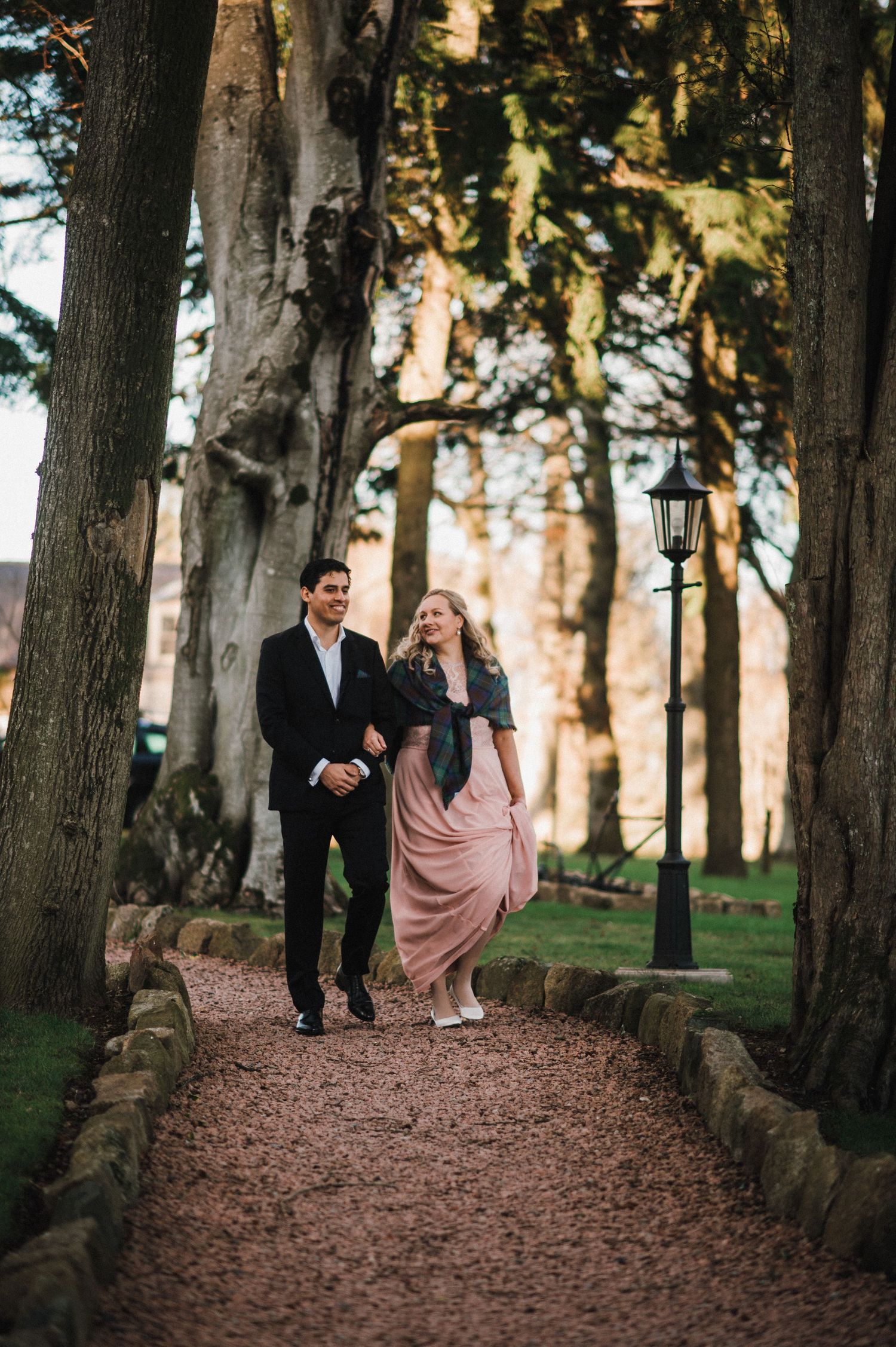A couple strolls down a tree-lined path with a vintage lamp post in the background.