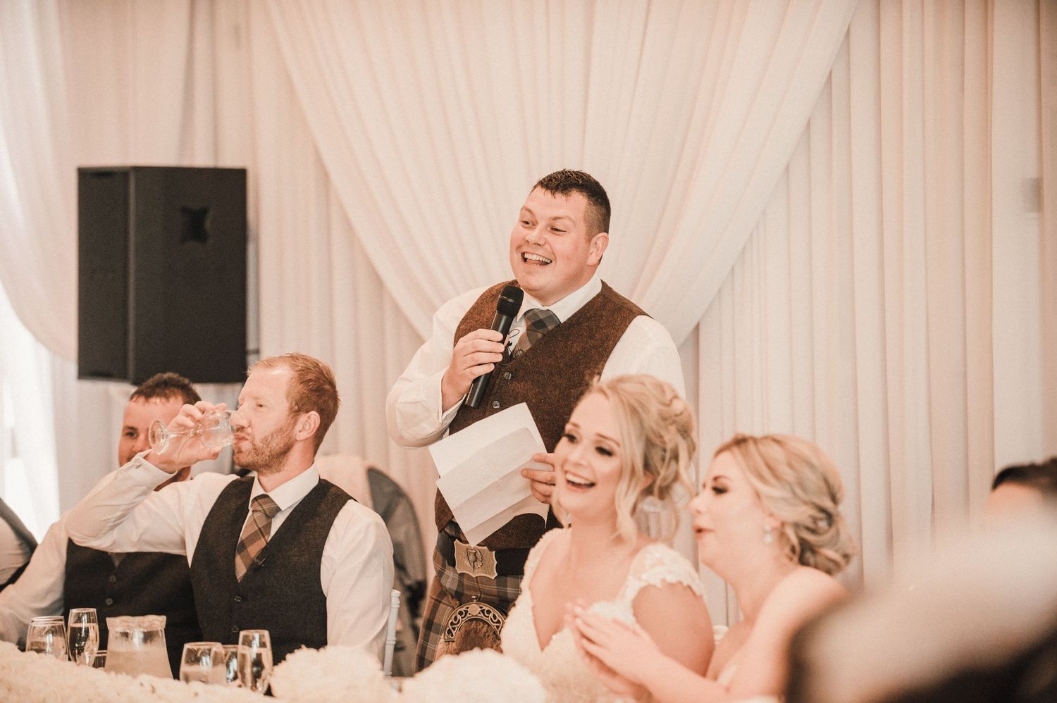 Wedding reception speech moment captured with guests seated at decorated tables.