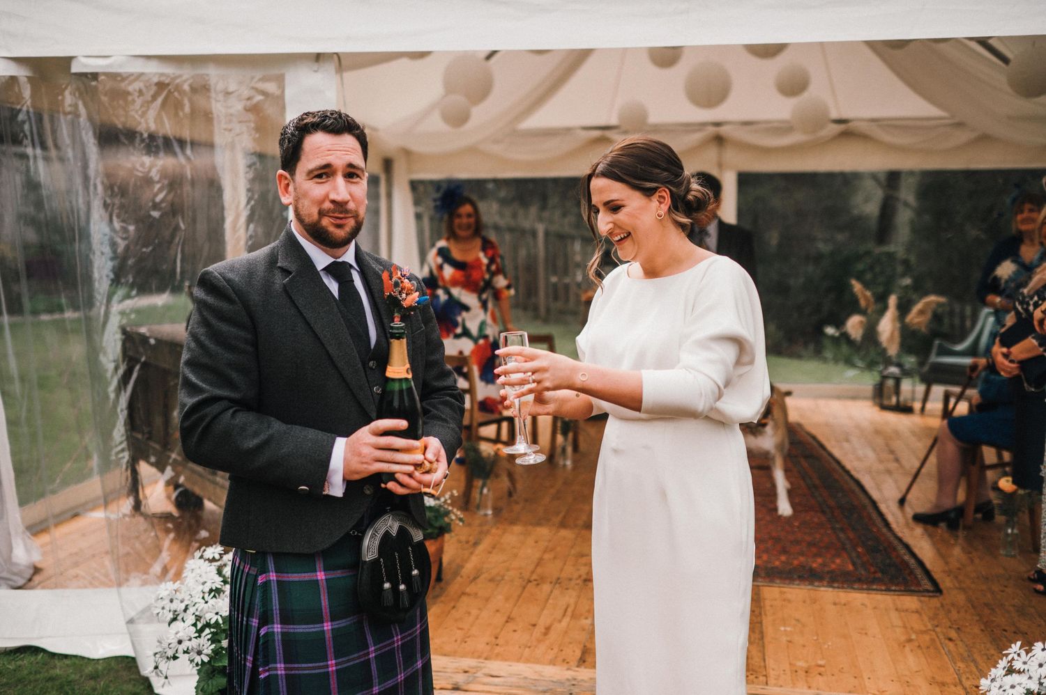 Wedding couple shares drinks during ceremony under white tent with wooden floor.