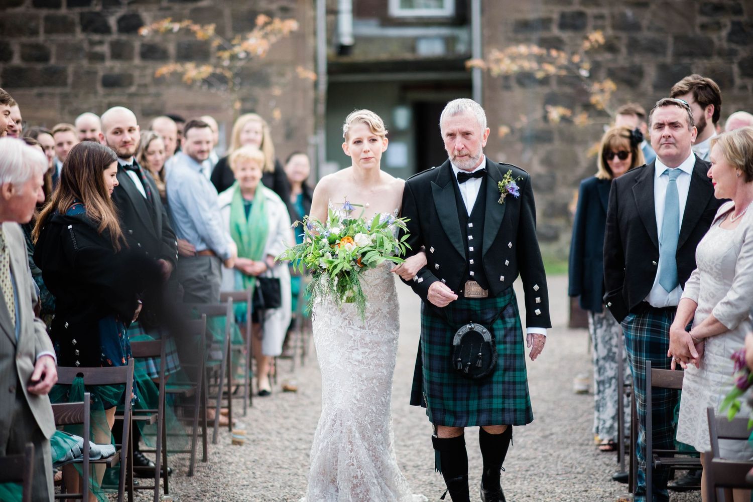 A Scottish wedding ceremony with attendees lining the path as a bride and gentleman in a traditional kilt walk down the aisle.