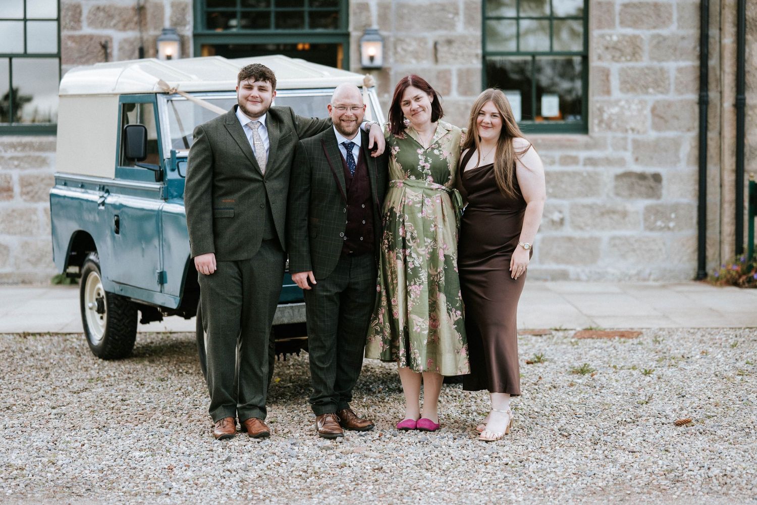 Group photo with vintage blue Land Rover truck against stone building backdrop.