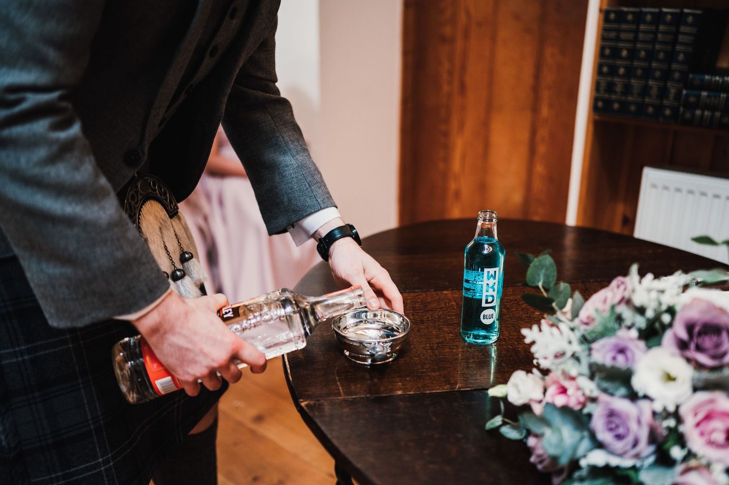 Wedding reception details showing hands pouring drinks at a decorated table with purple flowers.