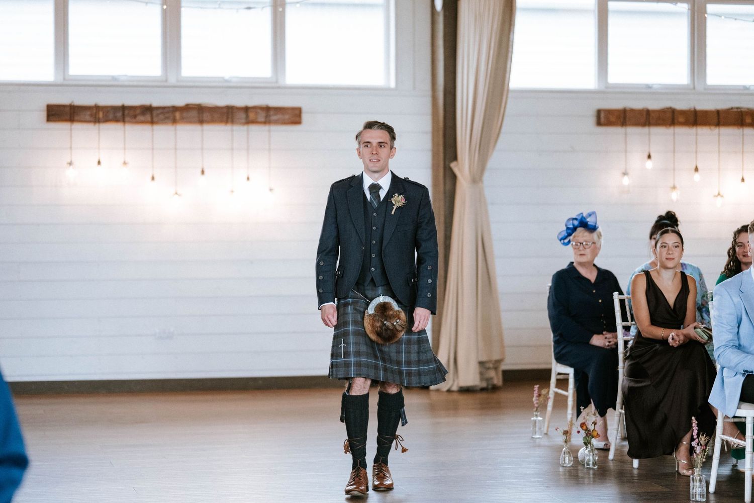 Wedding party members walking through an elegant venue space with large windows and hanging lights.