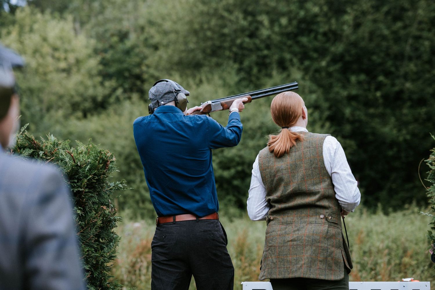 Two people participate in clay pigeon shooting at an outdoor range with one aiming a shotgun.