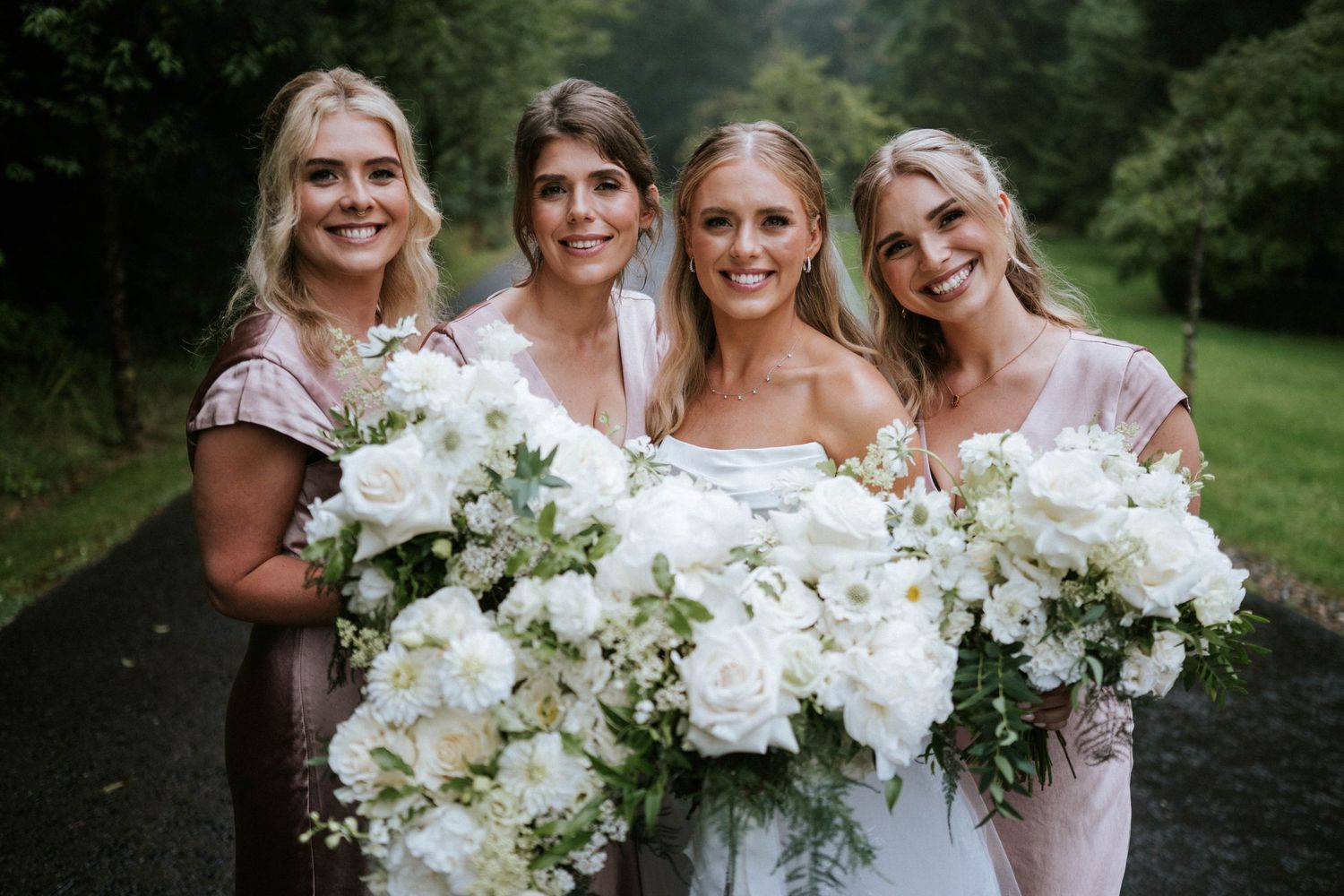 Bridesmaids in light dresses hold stunning white floral bouquets outdoors.