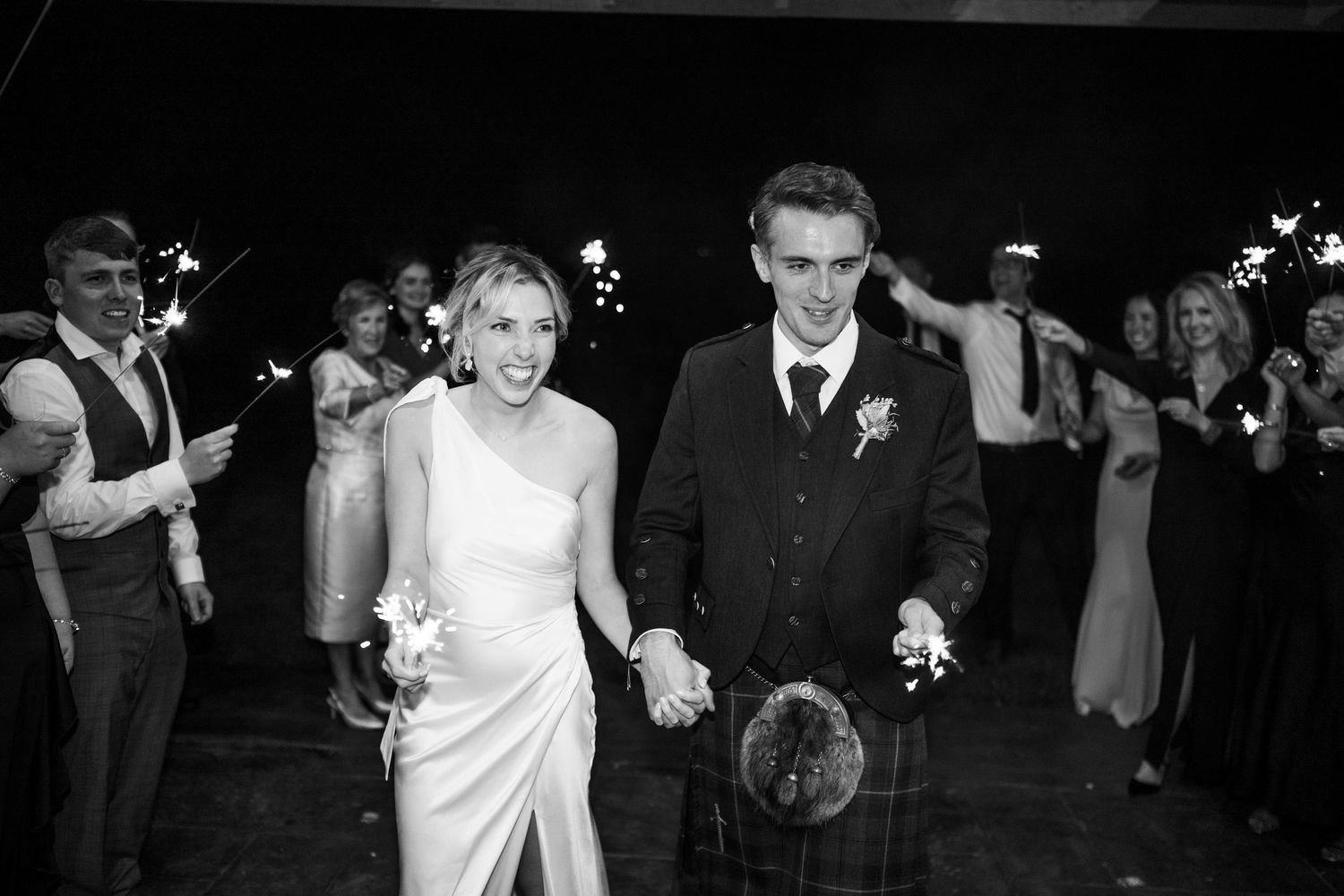 Black and white photo of newlyweds holding hands during sparkler exit.