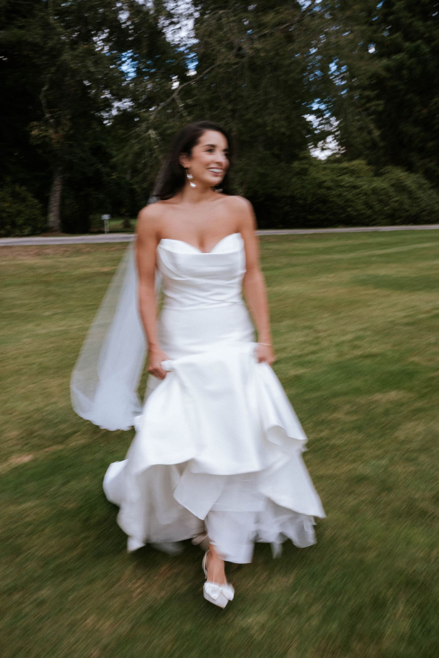 Bride in flowing strapless wedding gown walks gracefully across garden lawn with veil flowing.