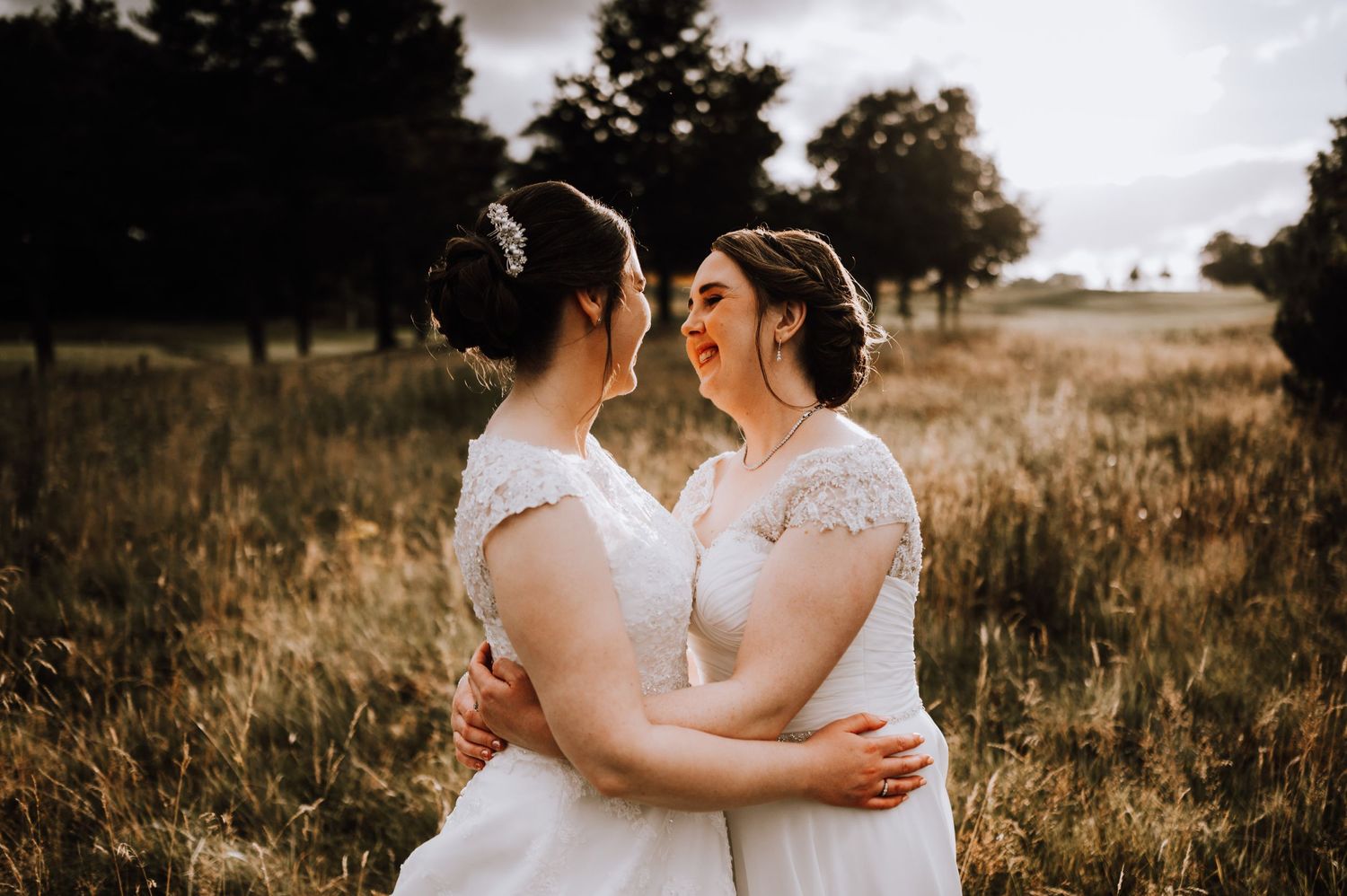 Romantic couple in matching white wedding dresses embracing in a golden field.