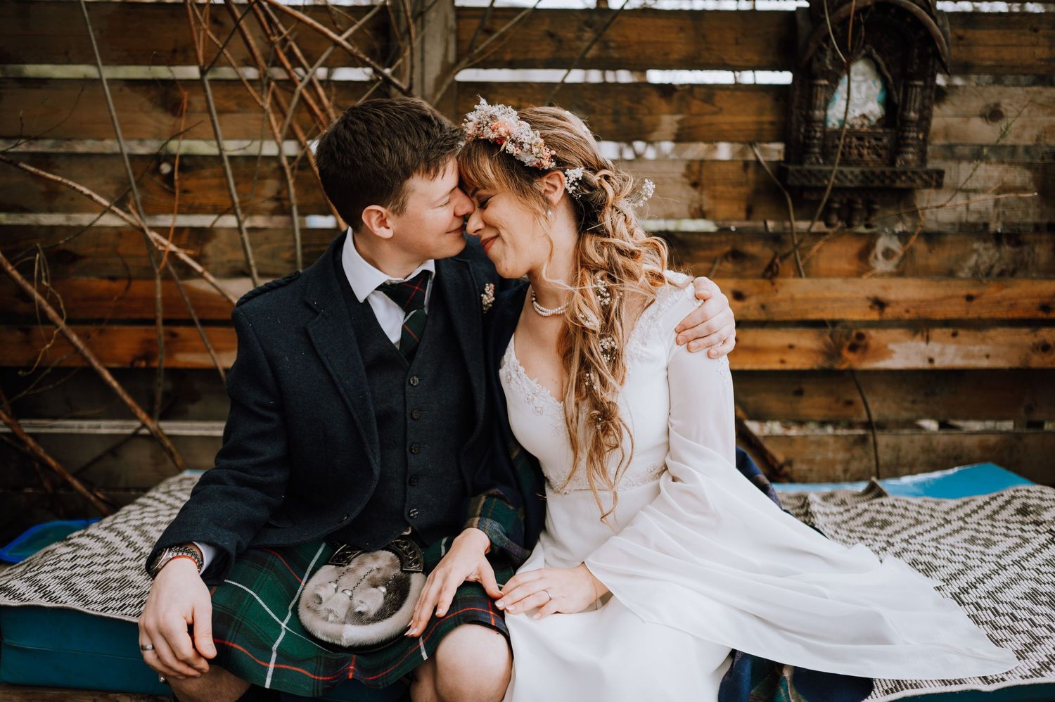 A romantic Scottish wedding couple sits intimately together, with the groom in a kilt and bride in long-sleeved white dress.