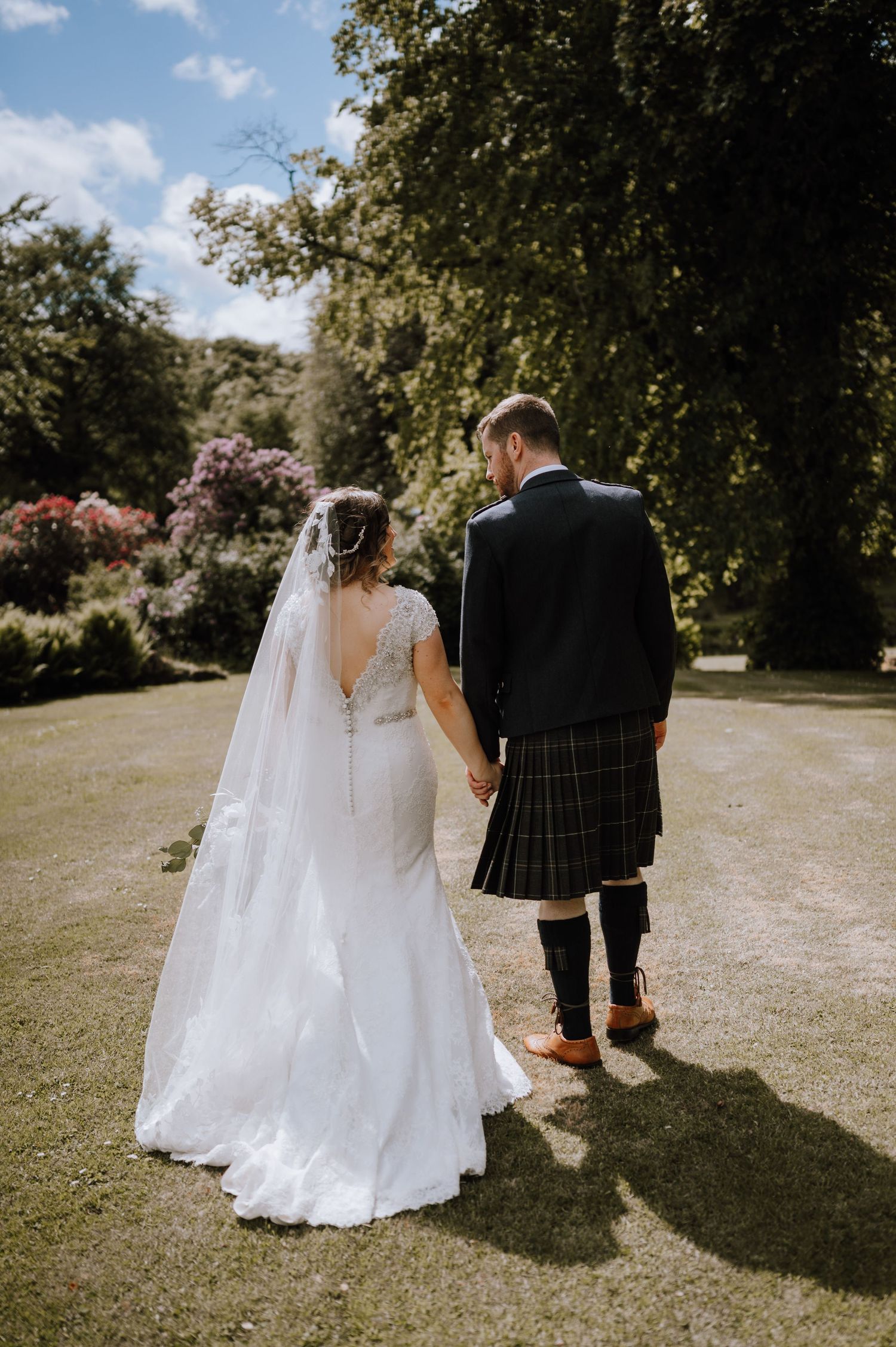 Newlyweds walking together on garden path with groom in traditional Scottish kilt.