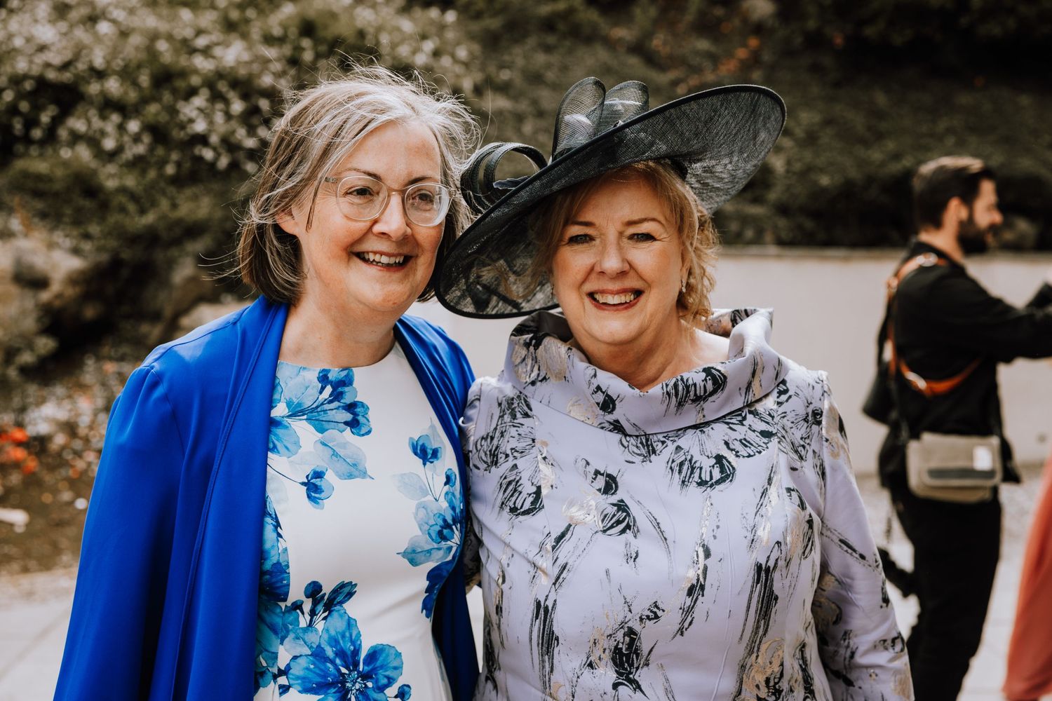 Two wedding guests in elegant dresses and hats smile together at an outdoor event.