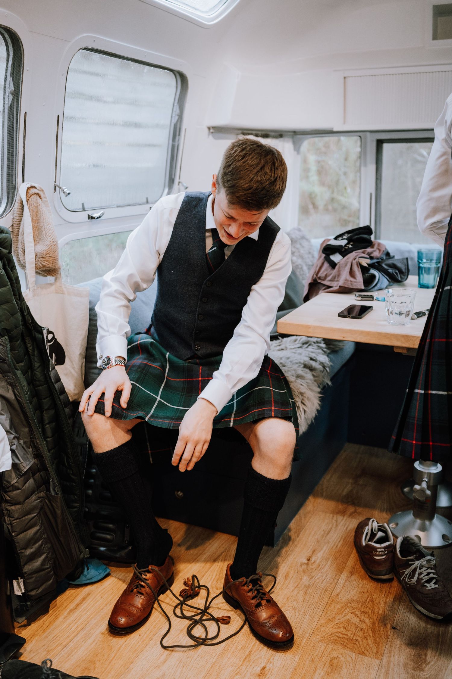 Person in traditional kilt and vest sitting in what appears to be an RV or mobile dressing room.